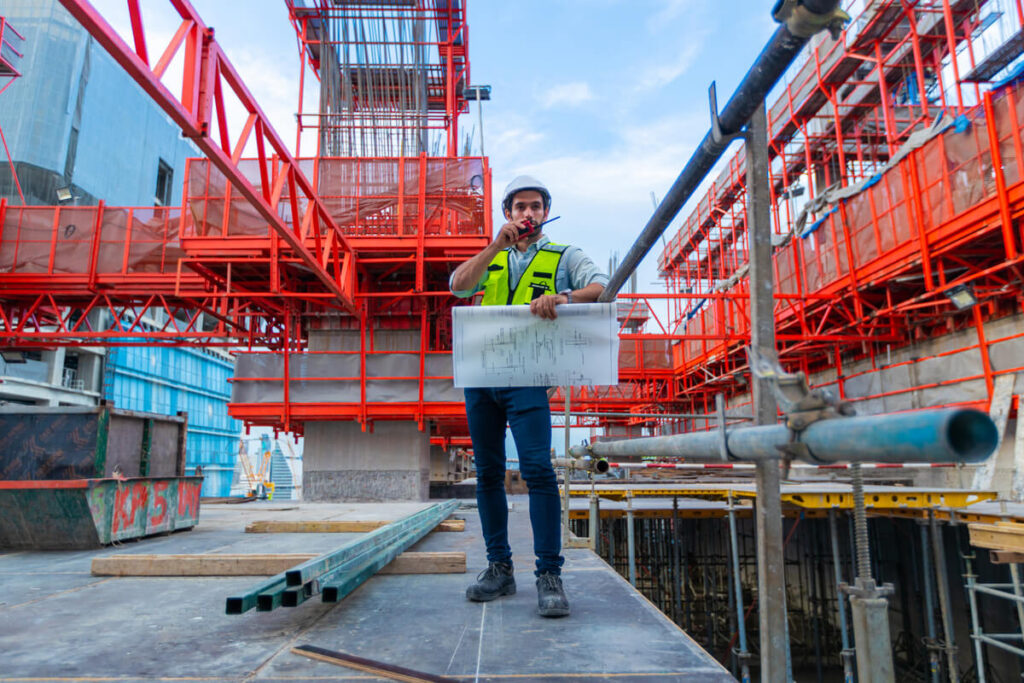 Quel artisan BTP a besoin d'une garantie décennale en auto-entrepreneur ? 2 Portrait d'un ingénieur en génie civil souriant, tenant les plans d'exécution d'un plancher en béton pour un chantier de construction.